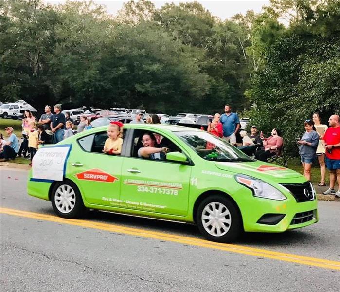 A green car is on a gray road surrounded by people.